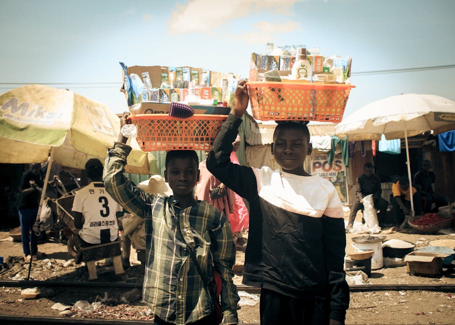 Two young street vendors carrying baskets on heads at a busy outdoor market.